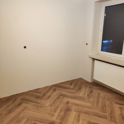DSK OPTIMA SP. Z O. O. - Pokój po remoncie: parkiet ułożony w jodełkę, białe ściany z widocznymi otworami na gniazdka, grzejnik pod oknem.