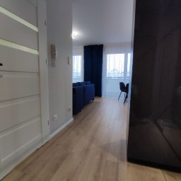 DSK OPTIMA SP. Z O. O. - Jasne panele podłogowe w nowocześnie urządzonym salonie z granatową sofą i zasłonami oraz białymi drzwiami.