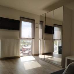DSK OPTIMA SP. Z O. O. - Jasne wnętrze pokoju z drewnianą podłogą, grzejnikami pod oknami z widokiem na miasto, telewizor na ścianie i szafą z lustrzanymi drzwiami, odbijającą fragment sofy.