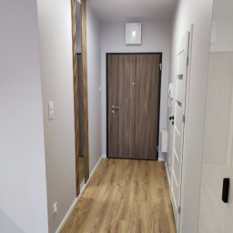 DSK OPTIMA SP. Z O. O. - Wąski korytarz po remoncie z panelami podłogowymi w odcieniach brązu, drewnianą dekoracją ścienną z pionowych listew, szarą ścianą i białymi drzwiami.