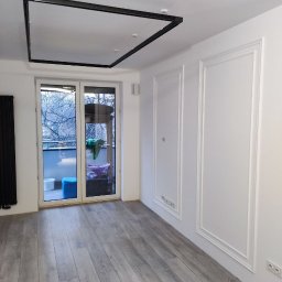 DSK OPTIMA SP. Z O. O. - Jasne wnętrze po remoncie: szare panele podłogowe, białe ściany z dekoracyjnymi listwami, czarna lampa sufitowa i przeszklone drzwi balkonowe.