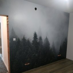 DSK OPTIMA SP. Z O. O. - Fototapeta z motywem mglistego lasu iglastego na ścianie w trakcie wykańczania, widoczne gniazdka elektryczne i fragment niedokończonego ościeża drzwiowego.