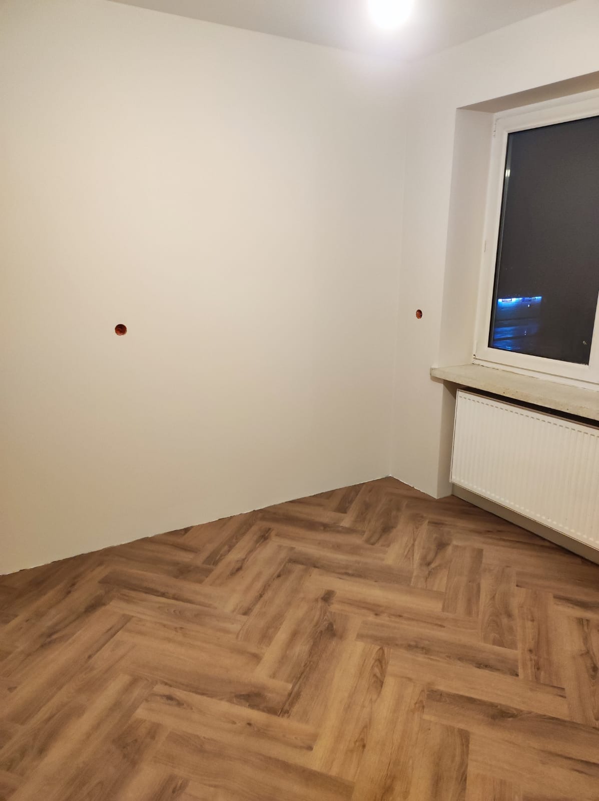 Pokój po remoncie: parkiet ułożony w jodełkę, białe ściany z widocznymi otworami na gniazdka, grzejnik pod oknem.