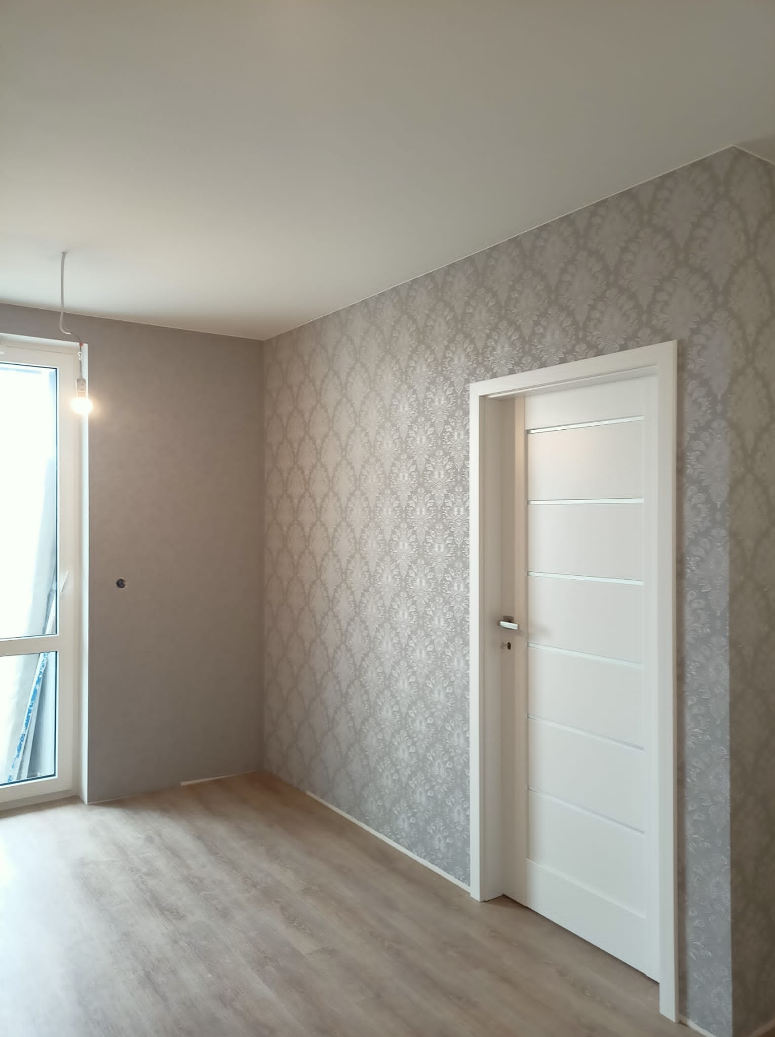 Pokój po remoncie: ściana pokryta tapetą w delikatny wzór, białe drzwi wewnętrzne z poziomymi wgłębieniami, panele podłogowe w odcieniach drewna, surowa żarówka zwisająca z sufitu.