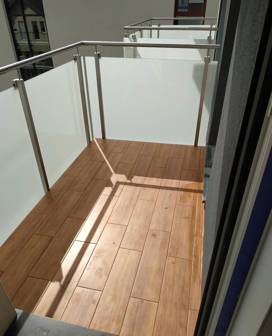 Balkon z drewnopodobnymi płytkami gresowymi, szklane balustrady z metalowymi ramami, cień barierki na podłodze.