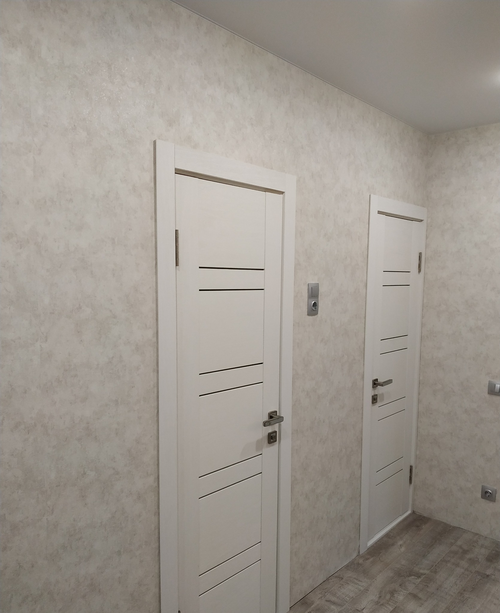 Wykończone wnętrze: Dwoje białych drzwi z poziomymi frezami na tle ściany z dekoracyjną fakturą i drewnopodobnej podłogi.