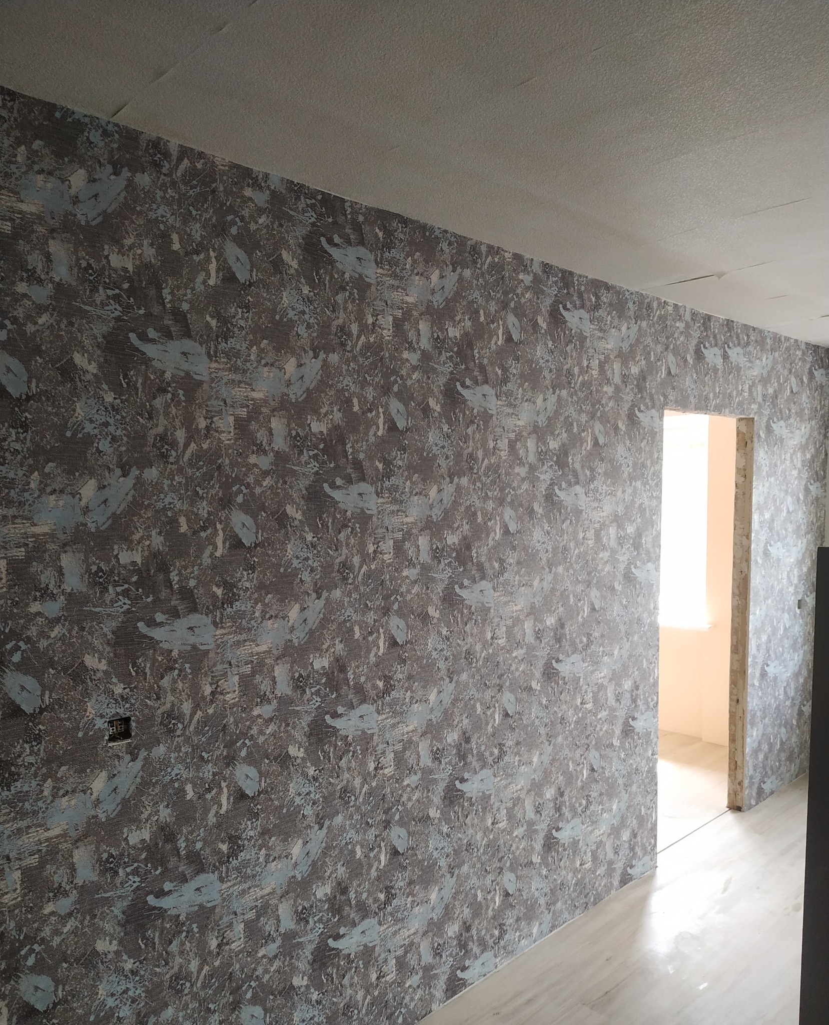 Ściana pokryta wzorzystą tapetą w odcieniach szarości i błękitu, widoczny fragment podłogi i sufitu, w tle otwór drzwiowy prowadzący do jasnego pomieszczenia.