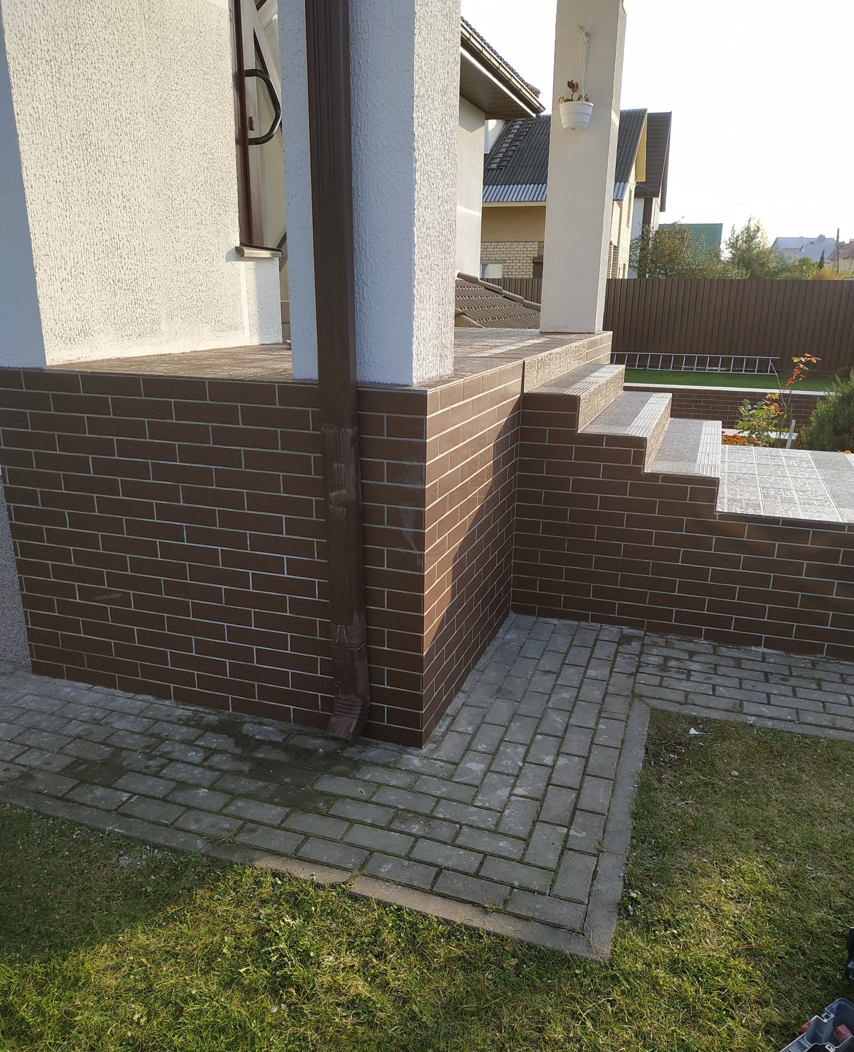 Wykończony taras z brązowych płytek imitujących cegłę, z dopasowanymi schodami i kolumną, brązowa rynna z boku, kostka brukowa i trawnik.