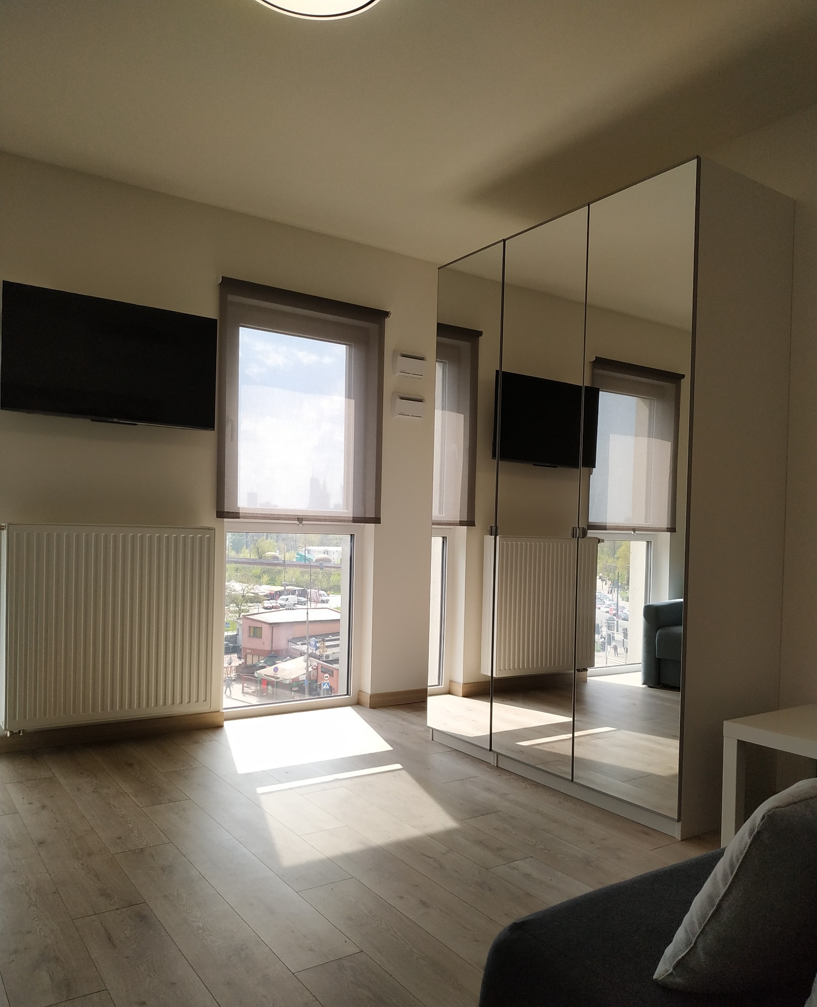 Jasne wnętrze pokoju z drewnianą podłogą, grzejnikami pod oknami z widokiem na miasto, telewizor na ścianie i szafą z lustrzanymi drzwiami, odbijającą fragment sofy.