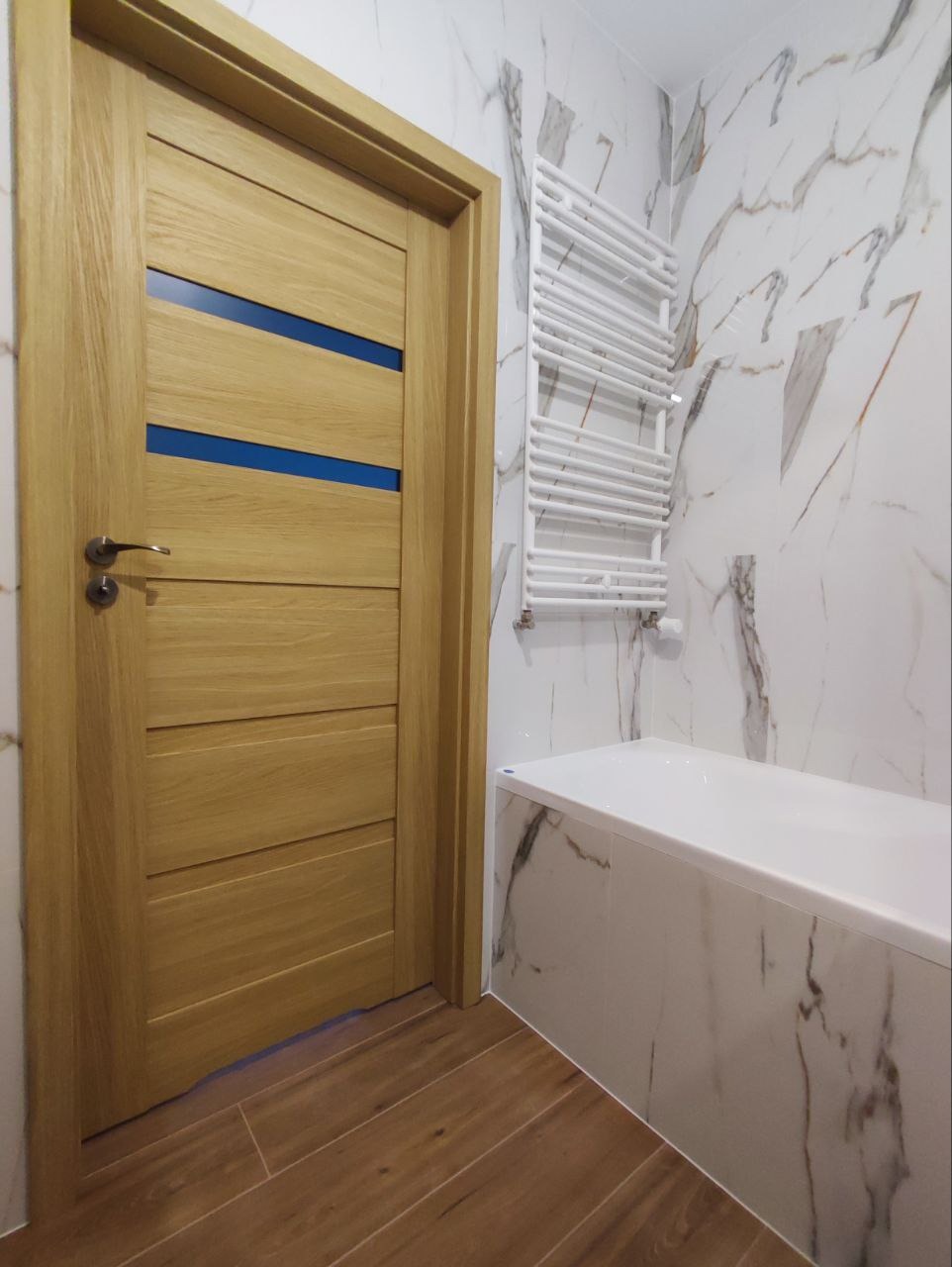 Wykończona łazienka z drewnianą podłogą, białą wanną obudowaną marmurowymi płytkami, grzejnikiem drabinkowym i drewnianymi drzwiami z niebieskimi wstawkami.