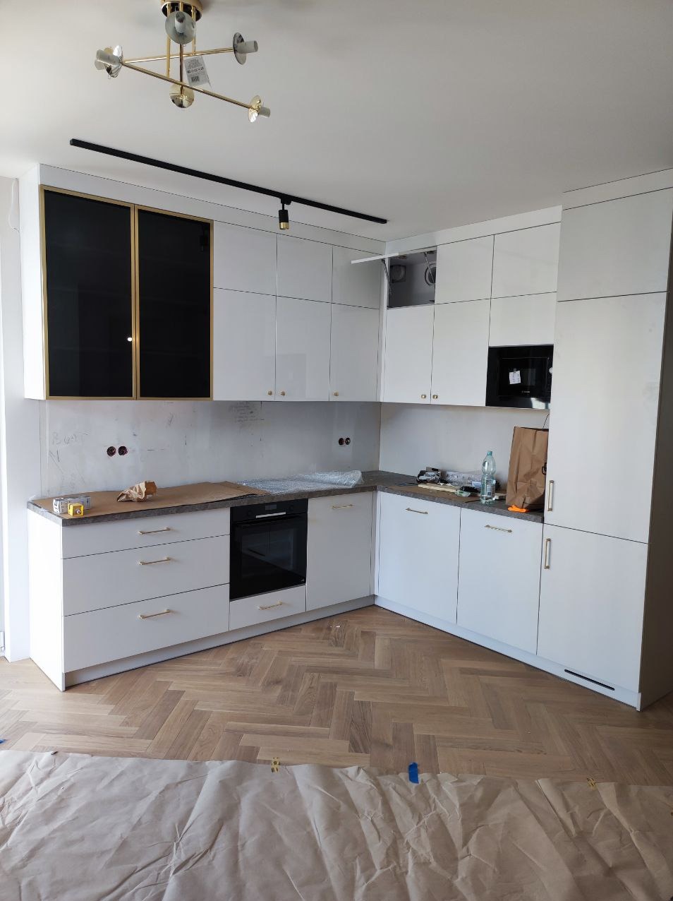Montaż białej, narożnej kuchni z czarnymi, lakierowanymi frontami górnych szafek, parkiet w jodełkę zabezpieczony papierem malarskim, widoczne ślady po instalacji elektrycznej na ścianie.