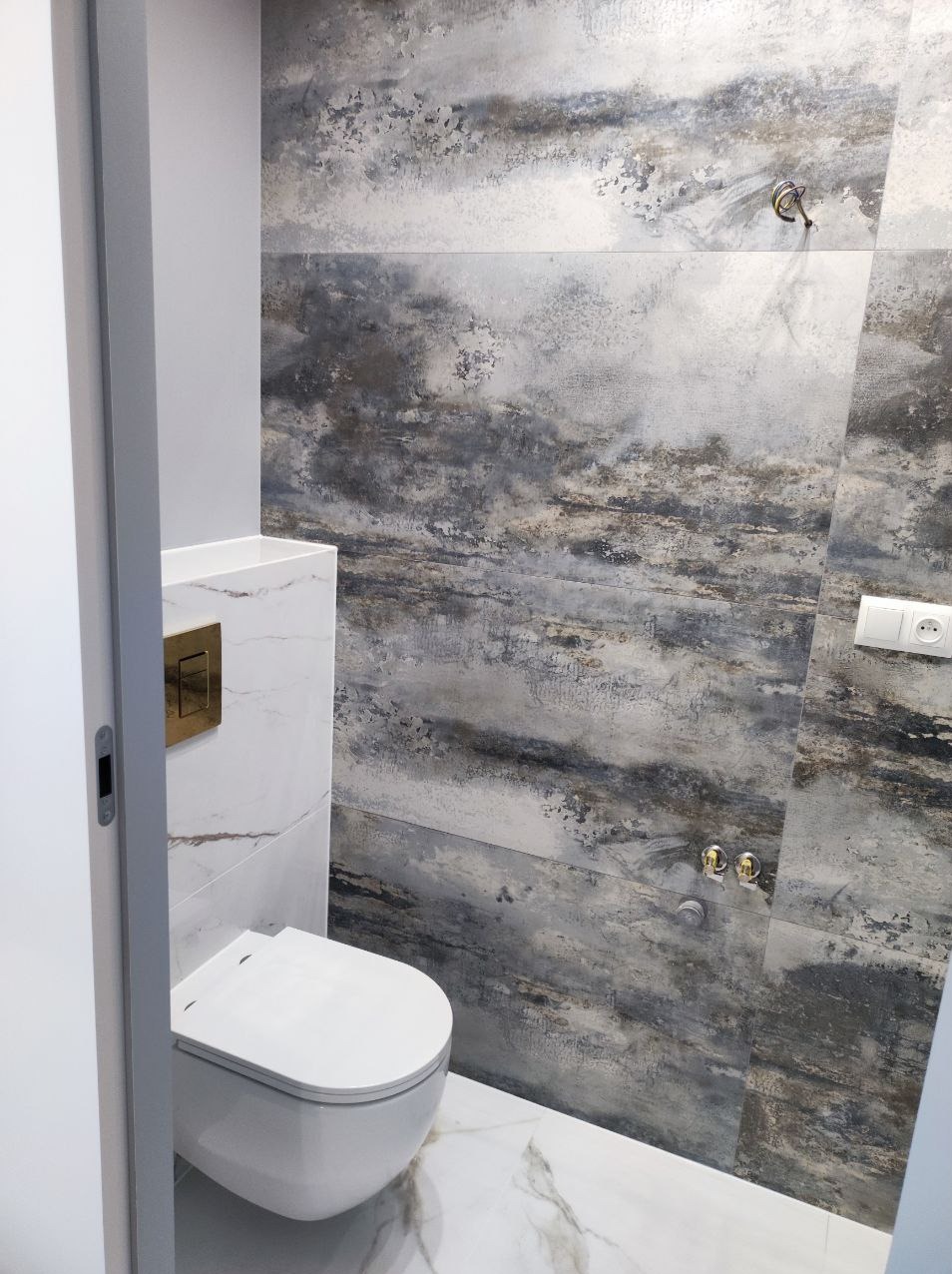 Nowoczesna toaleta w łazience wykończonej płytkami imitującymi beton, z marmurową podłogą i złotym przyciskiem spłukującym.