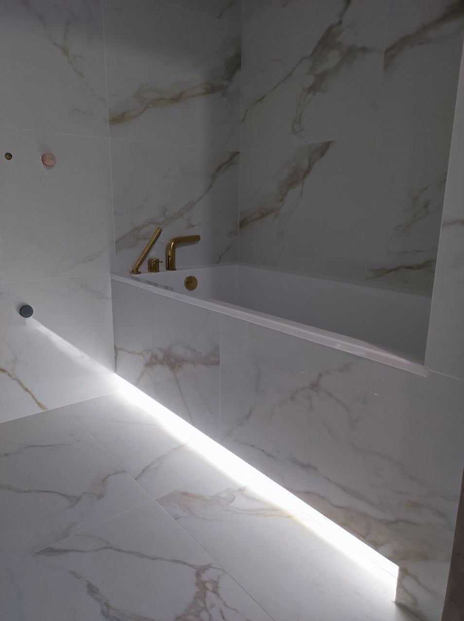 Nowoczesna łazienka z wanną obudowaną płytkami imitującymi marmur, oświetlenie LED pod wanną, złota armatura, widok z góry.