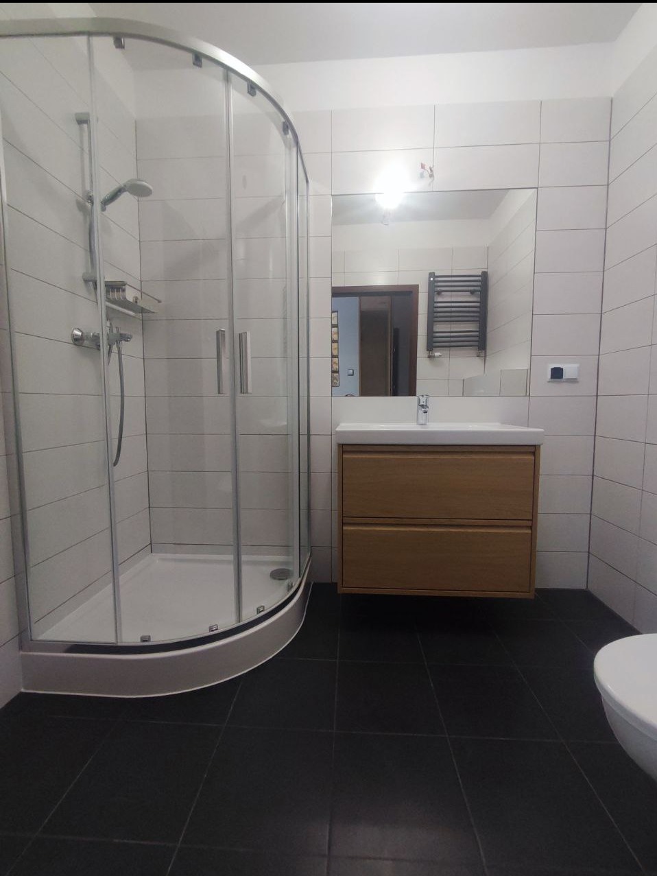 Wykończona łazienka z kabiną prysznicową z zaokrąglonym szkłem, umywalką z szafką z jasnego drewna i czarną podłogą z płytek.