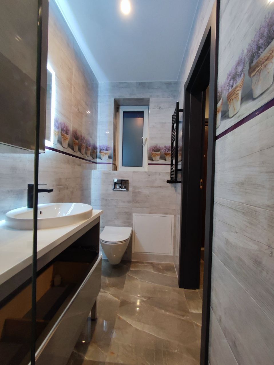 Nowoczesna łazienka z szarymi płytkami imitującymi drewno, dekoracyjnym motywem lawendy, podświetlanym lustrem nad umywalką i grafitowymi akcentami.