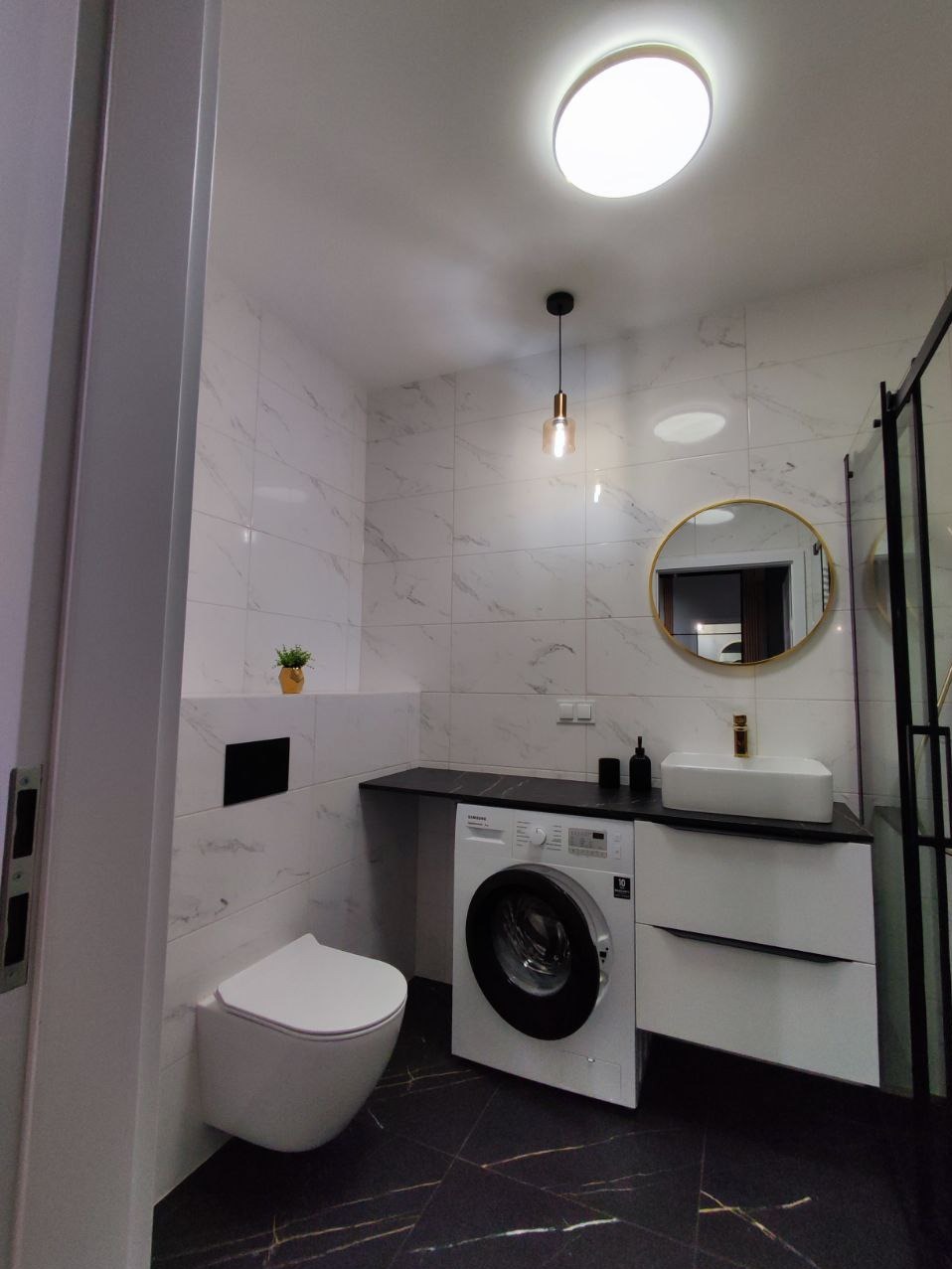 Nowoczesna łazienka w Warszawie z białymi płytkami imitującymi marmur, czarną podłogą z złotymi żyłkami, wiszącą toaletą, pralką pod blatem umywalkowym i okrągłym lustrem w złotej ramie.