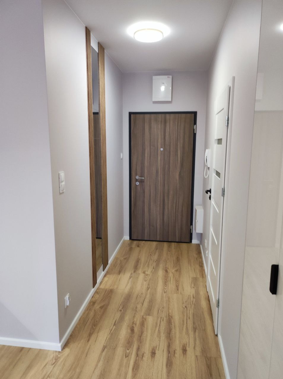 Wąski korytarz po remoncie z panelami podłogowymi w odcieniach brązu, drewnianą dekoracją ścienną z pionowych listew, szarą ścianą i białymi drzwiami.