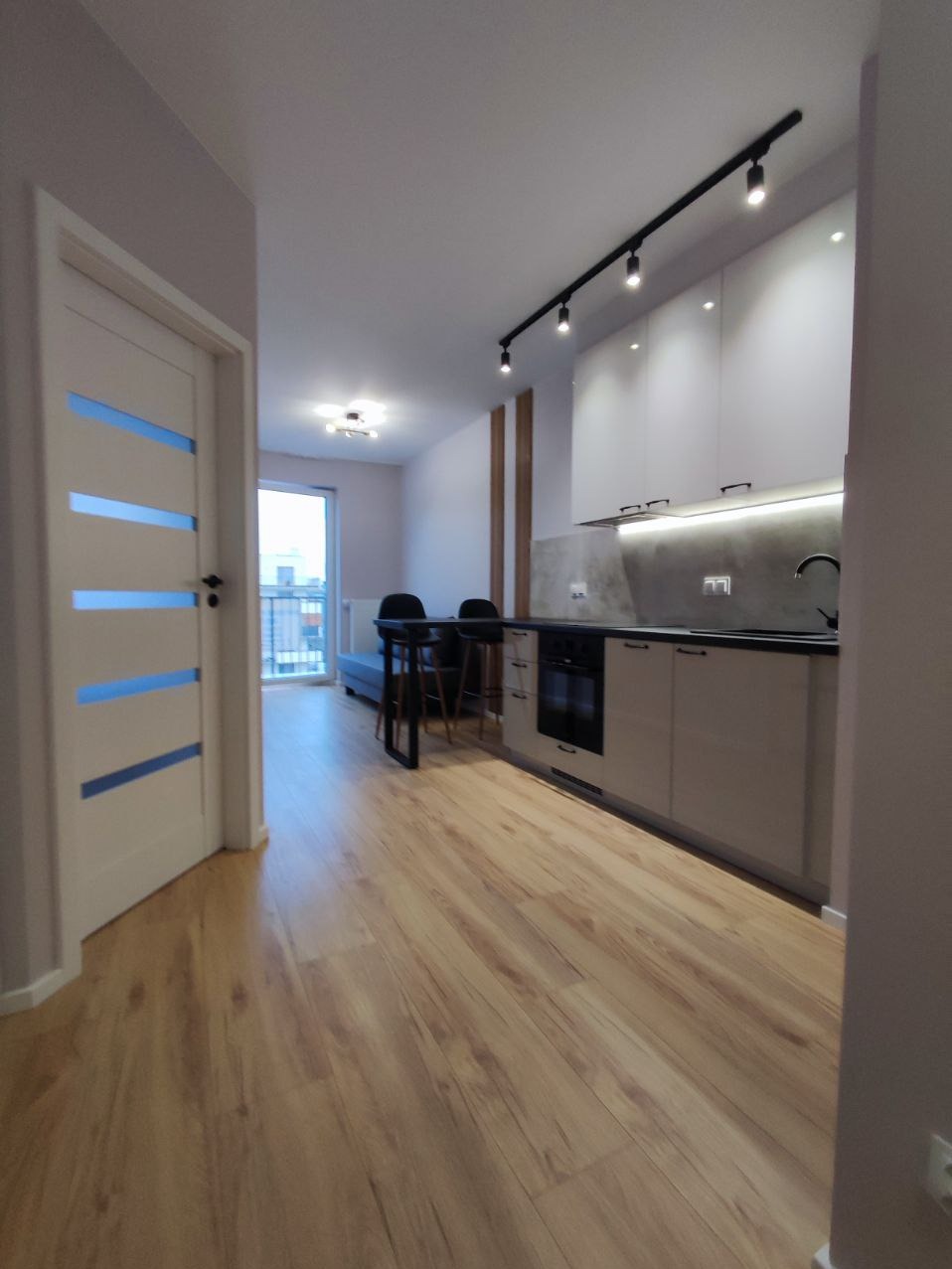 Nowoczesna kuchnia w jasnych barwach z drewnianą podłogą, czarnym oświetleniem punktowym i minimalistycznym stołem barowym z krzesłami w tle balkonu.