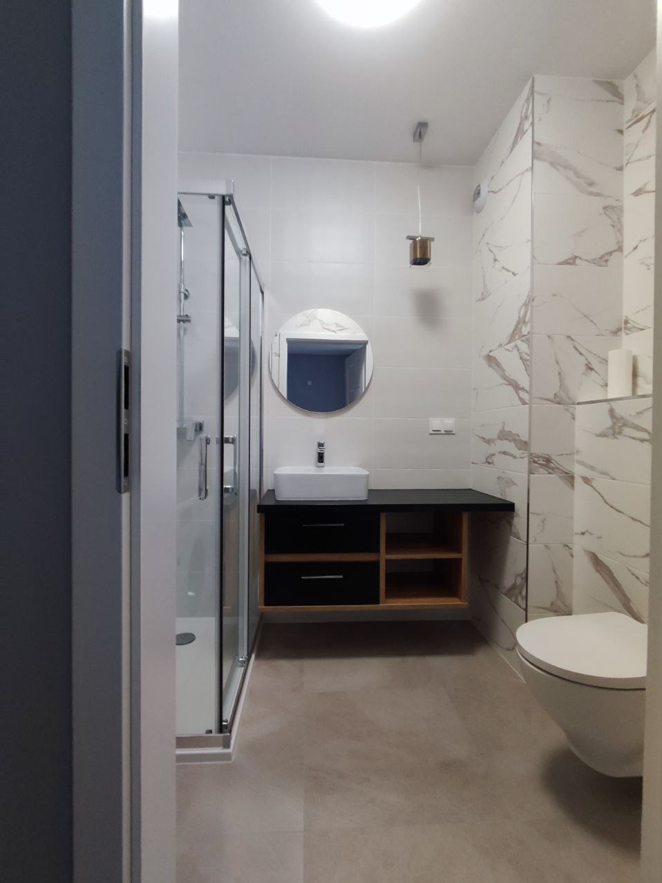 Wykończona łazienka z kabiną prysznicową, umywalką na szafce z czarnymi szufladami i półkami z jasnego drewna, okrągłym lustrem i toaletą, płytki imitujące marmur na ścianie za toaletą, sufitowe...