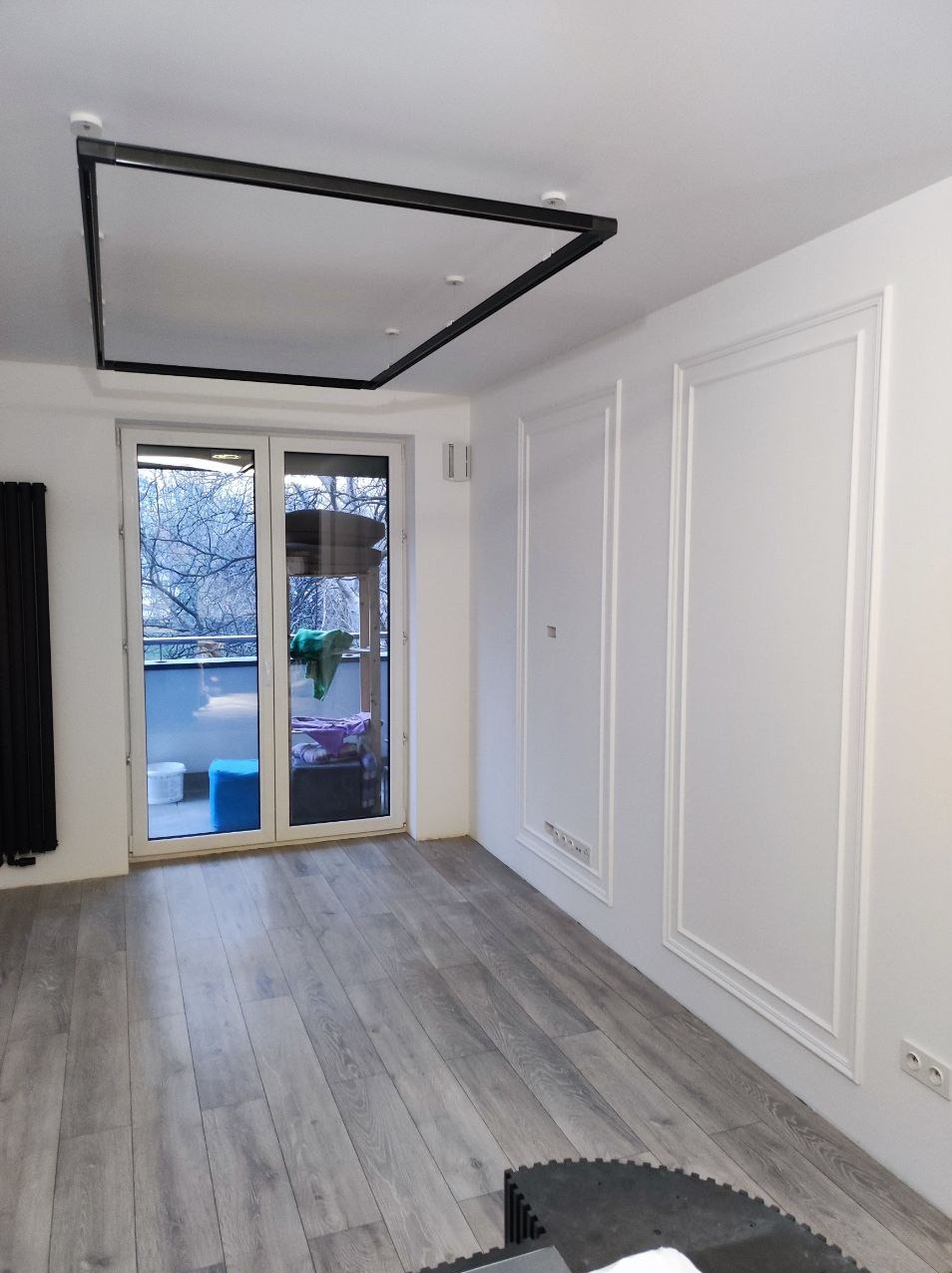 Jasne wnętrze po remoncie: szare panele podłogowe, białe ściany z dekoracyjnymi listwami, czarna lampa sufitowa i przeszklone drzwi balkonowe.