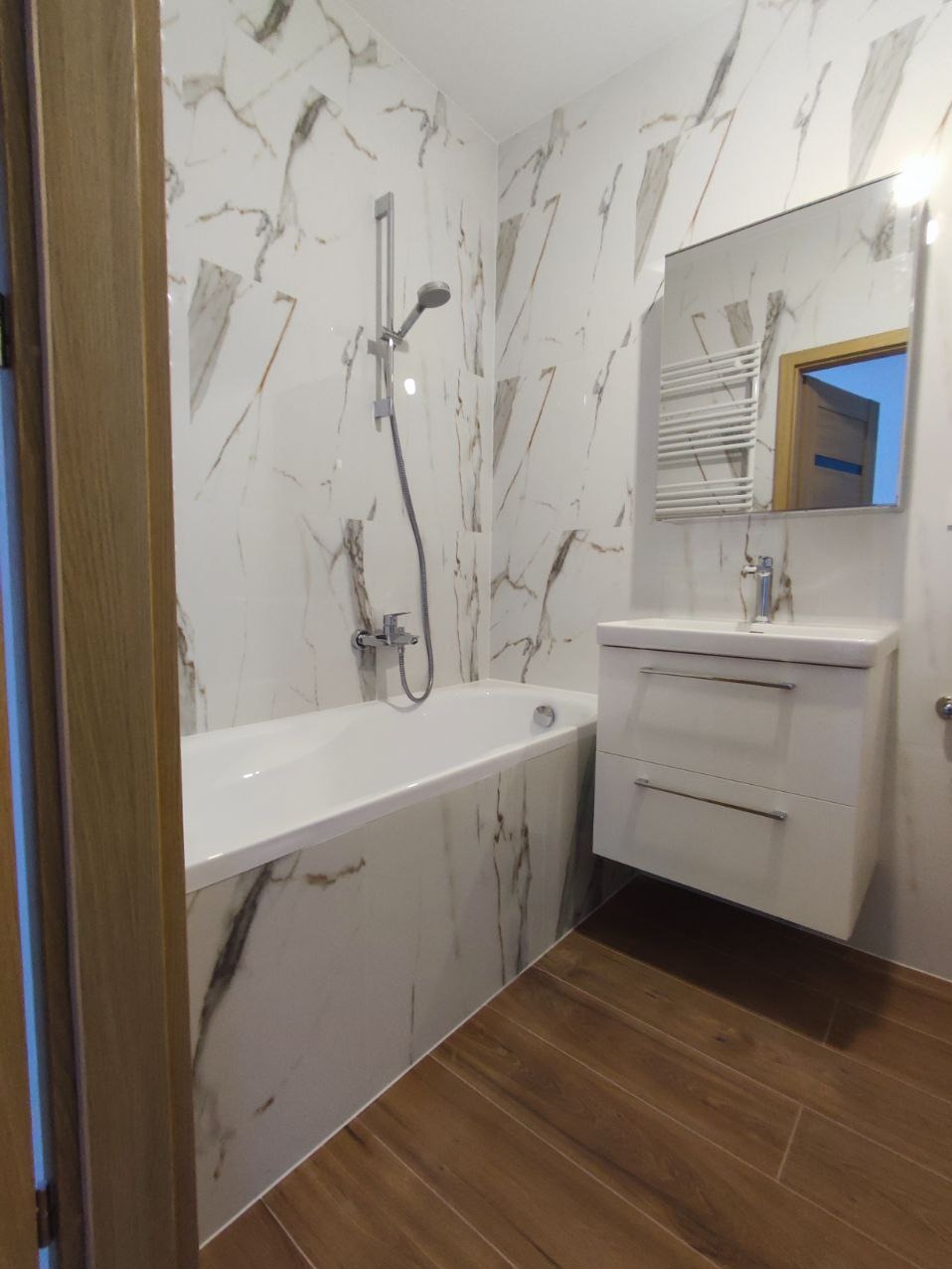 Biała wanna obudowana płytkami imitującymi marmur w łazience, z baterią prysznicową i umywalką z szafką w tle oraz drewnopodobną podłogą.