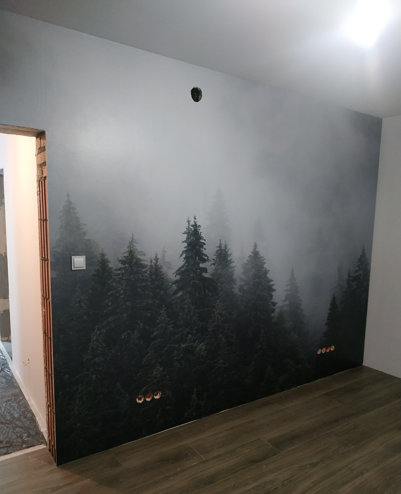 Fototapeta z motywem mglistego lasu iglastego na ścianie w trakcie wykańczania, widoczne gniazdka elektryczne i fragment niedokończonego ościeża drzwiowego.