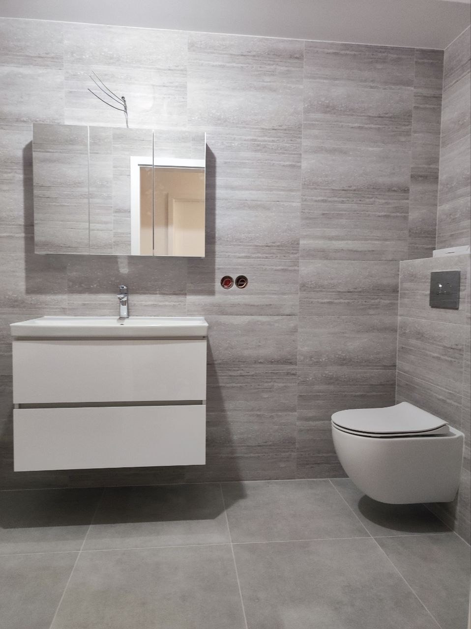 Nowoczesna łazienka w odcieniach szarości z podwieszaną szafką umywalkową, lustrem z szafkami i wiszącą toaletą.