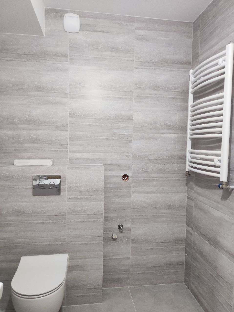 Wykończona łazienka z szarymi płytkami imitującymi drewno, wiszącą toaletą, chromowanym przyciskiem spłuczki, białym grzejnikiem drabinkowym i widocznymi przyłączami wodnymi.