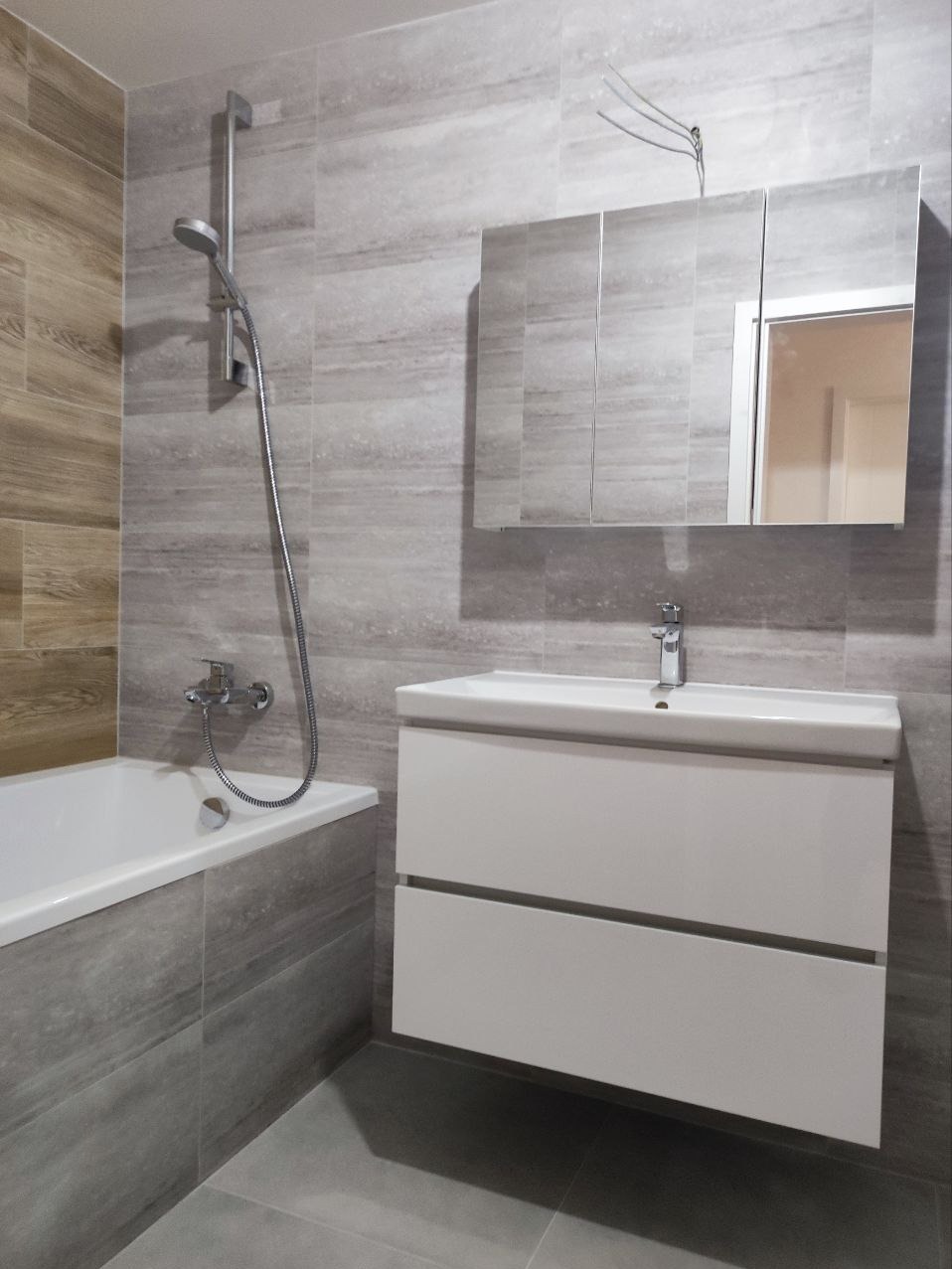 Nowoczesna łazienka z szarymi płytkami imitującymi kamień, białą szafką pod umywalką i lustrem z szafkami, widoczna bateria umywalkowa i słuchawka prysznicowa.
