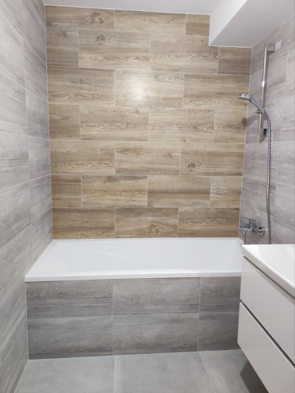 Nowoczesna łazienka z wanną obudowaną szarymi płytkami imitującymi kamień, ściana nad wanną wyłożona płytkami drewnopodobnymi ułożonymi poziomo, chromowana bateria i słuchawka prysznicowa.
