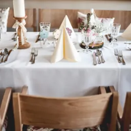 Elegancko nakryty stół z białym obrusem, świecą na drewnianym świeczniku z koronkową wstążką, szklankami, sztućcami, serwetami ułożonymi w trójkąty oraz dekoracjami kwiatowymi w małych wazonach...