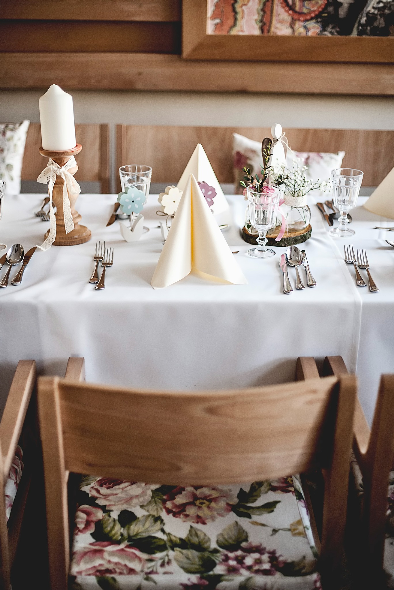 Elegancko nakryty stół z białym obrusem, świecą na drewnianym świeczniku z koronkową wstążką, szklankami, sztućcami, serwetami ułożonymi w trójkąty oraz dekoracjami kwiatowymi w małych wazonach...