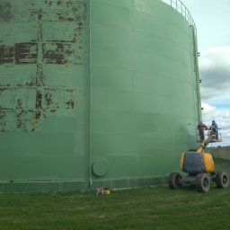 Profmalowanie.pl - Dwóch robotników na podnośniku koszowym odnawia zielony, przemysłowy zbiornik; widoczne ślady rdzy i odbarwienia na części powierzchni, świeża farba na pozostałej części.