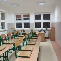 Profmalowanie.pl - Pomalowana klasa szkolna z rzędami ławek z krzesłami o zielonych ramach, jasnymi ścianami z białymi oknami i grzejnikiem, w tle biurko z odwróconym krzesłem.