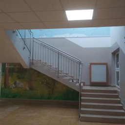 Profmalowanie.pl - Wnętrze szkoły z pomalowaną ścianą przedstawiającą las, schody z metalową balustradą, na górze widoczna mapa świata na ścianie.