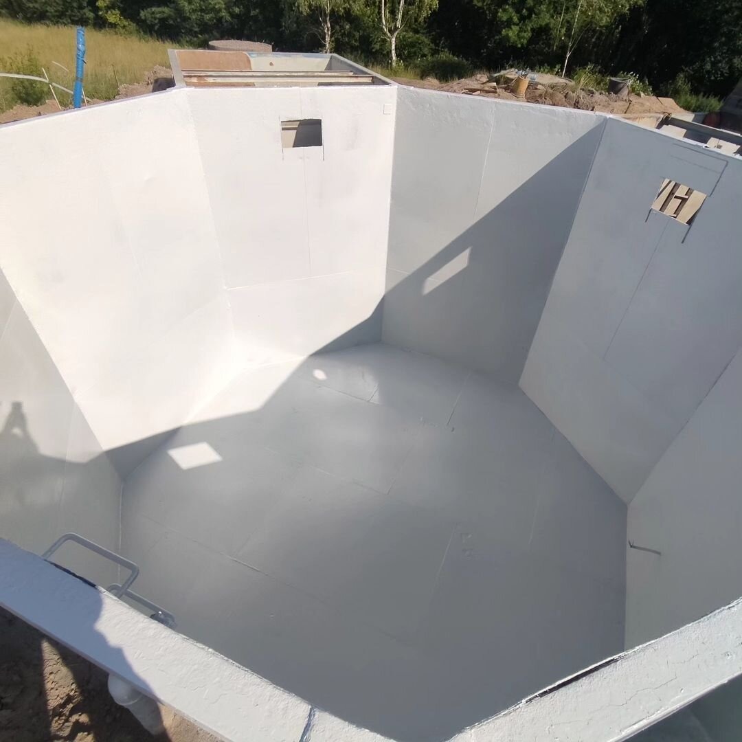 Wnętrze świeżo pomalowanego na biało zbiornika o nietypowym, wielokątnym kształcie, z widocznymi śladami łączeń płyt i niewielkimi otworami w ścianach, z drabinką u dołu i cieniem rzucanym od góry,...