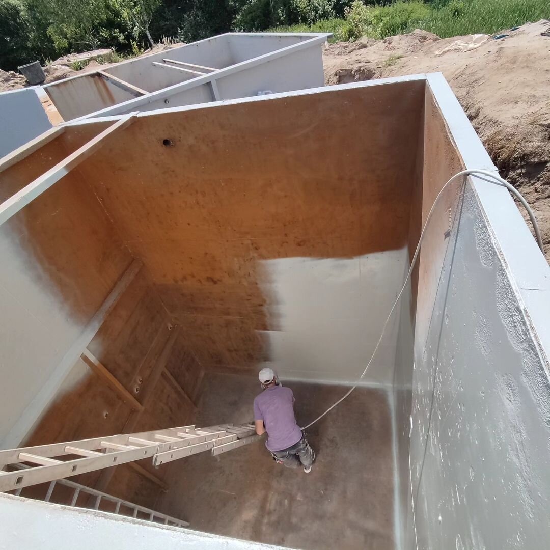 Pracownik w masce i kombinezonie maluje natryskowo wnętrze betonowego zbiornika na drabinie, widoczne częściowo pomalowane ściany i dno zbiornika.