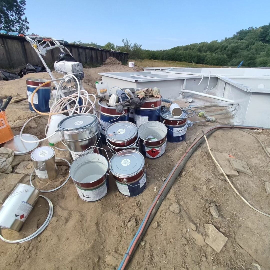 Przygotowanie do malowania zbiorników: widoczne puszki farb, sprzęt do malowania natryskowego i częściowo pomalowane zbiorniki na zewnątrz.