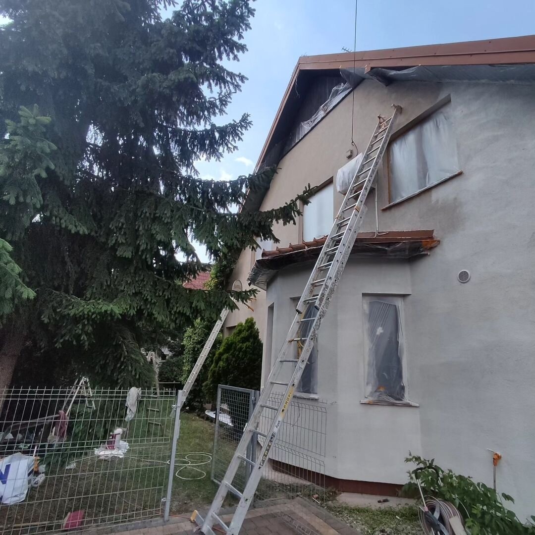 Malowanie elewacji domu jednorodzinnego: widoczna drabina oparta o ścianę, okna zabezpieczone folią, świeżo pomalowana, jasnoszara elewacja w trakcie prac wykończeniowych.