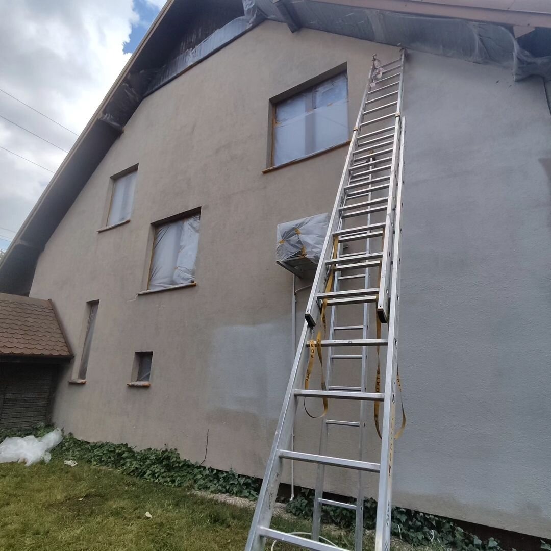 Dom jednorodzinny z elewacją w trakcie odświeżania, okna zabezpieczone folią, widoczna aluminiowa drabina oparta o ścianę, ślady świeżo nałożonej farby.