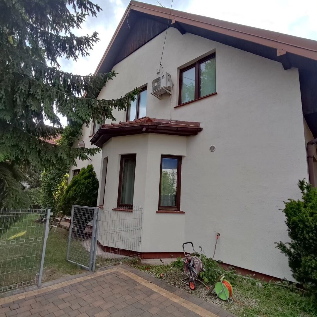 Jasna elewacja domu jednorodzinnego z brązowymi ramami okiennymi, widoczna jednostka zewnętrzna klimatyzacji, fragment ogrodu z siatką ogrodzeniową, wąż ogrodowy na wózku i zwinięty wąż na bębnie.