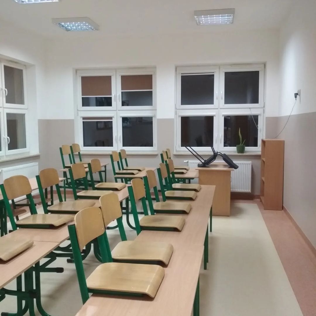 Pomalowana klasa szkolna z rzędami ławek z krzesłami o zielonych ramach, jasnymi ścianami z białymi oknami i grzejnikiem, w tle biurko z odwróconym krzesłem.