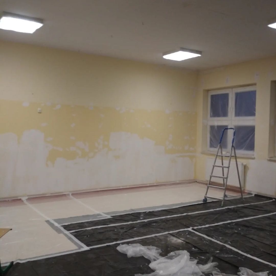 Przygotowanie sali szkolnej do odświeżenia: ściany częściowo zagruntowane, okna zabezpieczone folią, podłoga przykryta czarną folią ochronną, drabina przystawiona do okna.
