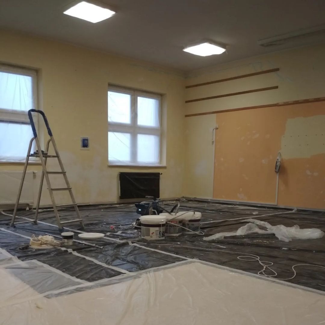 Przygotowane do malowania wnętrze szkoły: podłoga zabezpieczona folią, drabina oparta o ścianę, widoczne puszki z farbą i agregat malarski.