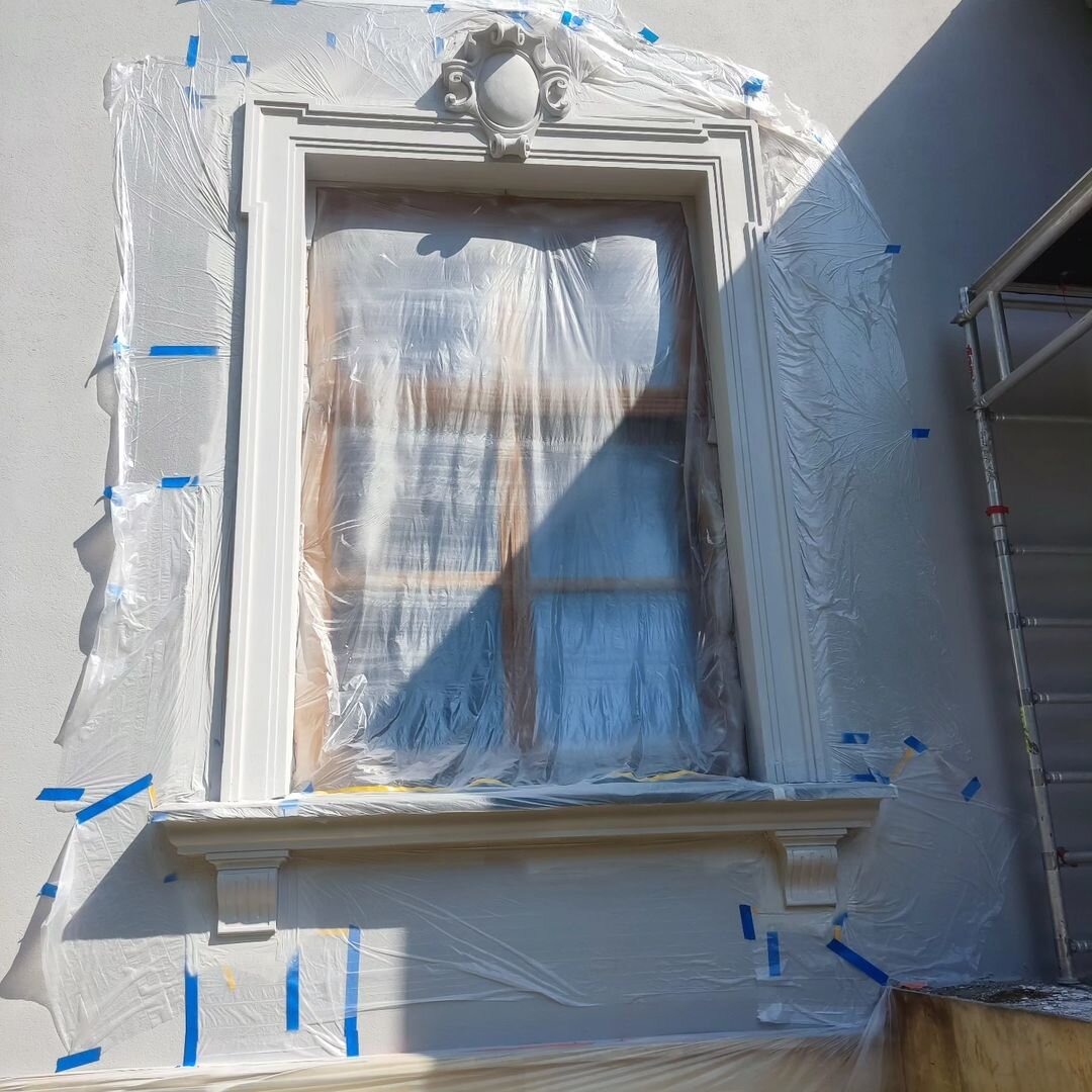 Okna z dekoracyjną sztukaterią elewacyjną zabezpieczone folią i taśmą malarską przed pracami malarskimi, rusztowanie obok.