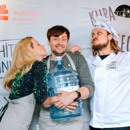 WYJĄTKOWY PREZENT SPÓŁKA Z OGRANICZONĄ ODPOWIEDZIALNOŚCIĄ - Trzy osoby w fartuchach pozują z dużym baniakiem wody i chochlą na tle ściany z napisem 'Wyjątkowy Prezent' i logo 'Shitty Dinner'.