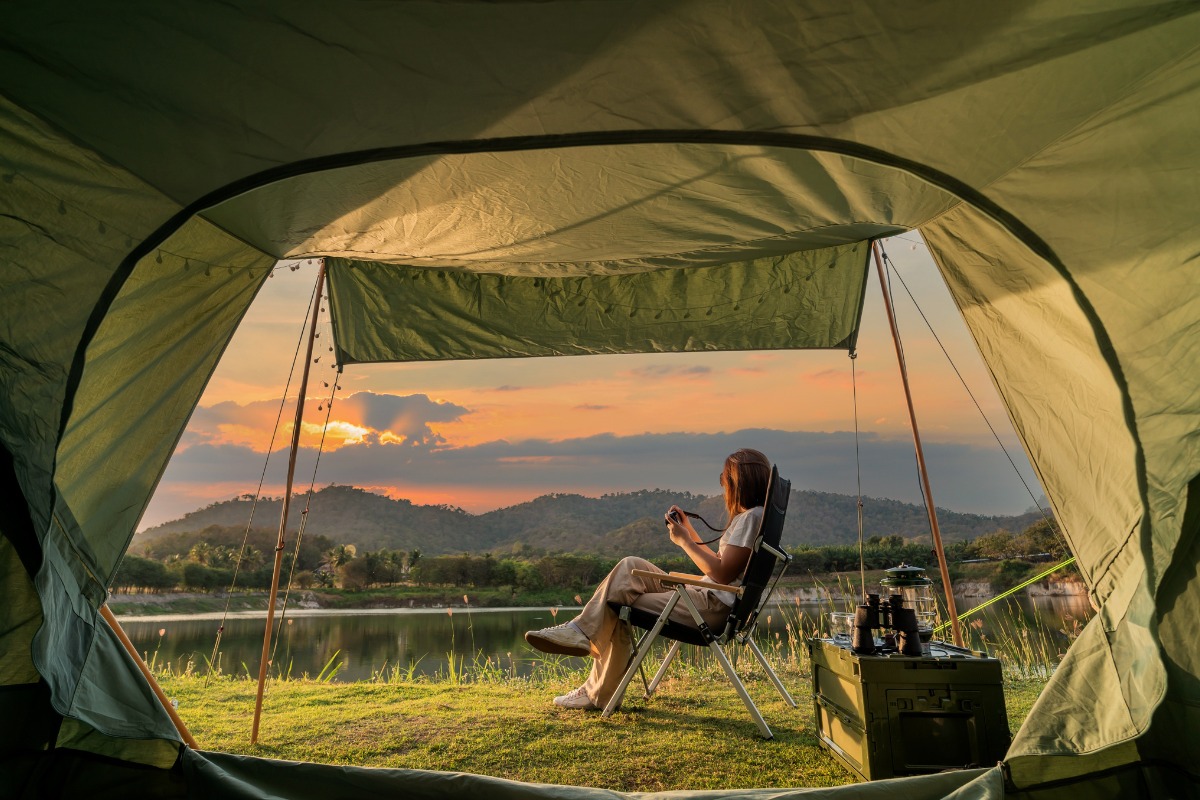 Kobieta siedzi w rozłożonym krześle turystycznym w namiocie, fotografując krajobraz z jeziorem i zachodzącym słońcem