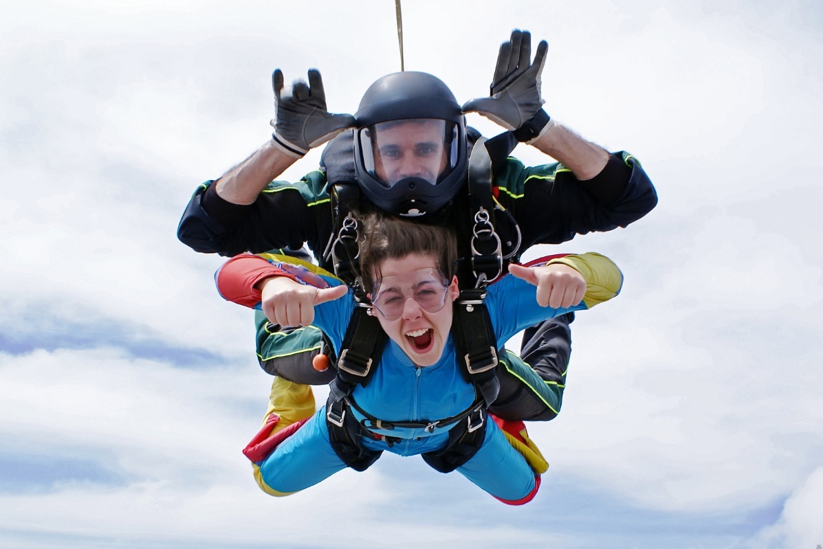 Tandemowy skok spadochronowy: instruktor w czarnym kasku z wizjerem i rękawicach pokazuje rogi, a podekscytowana kobieta w niebieskim kombinezonie i okularach ochronnych unosi kciuki w górę na tle...