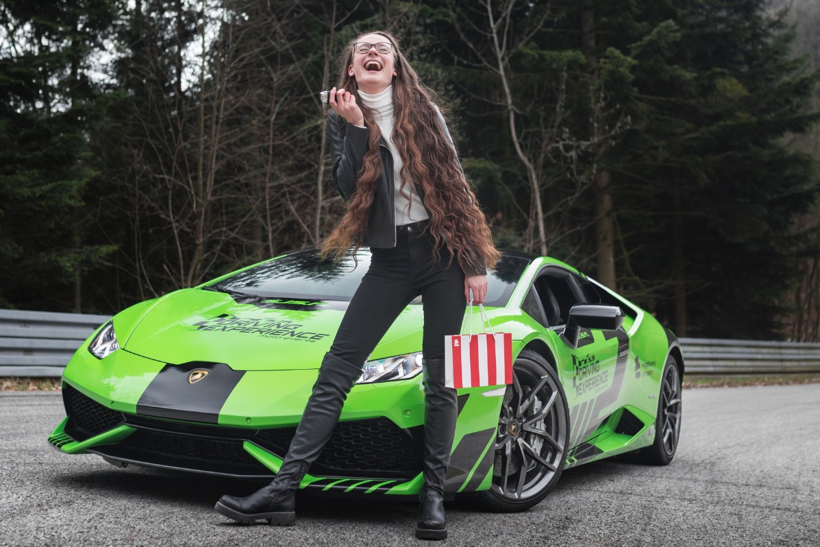 Uśmiechnięta kobieta w okularach, czarnej kurtce i kozakach, trzymająca torby na zakupy, stoi przed jaskrawozielonym Lamborghini z napisem 'Driving Experience' na tle lasu.