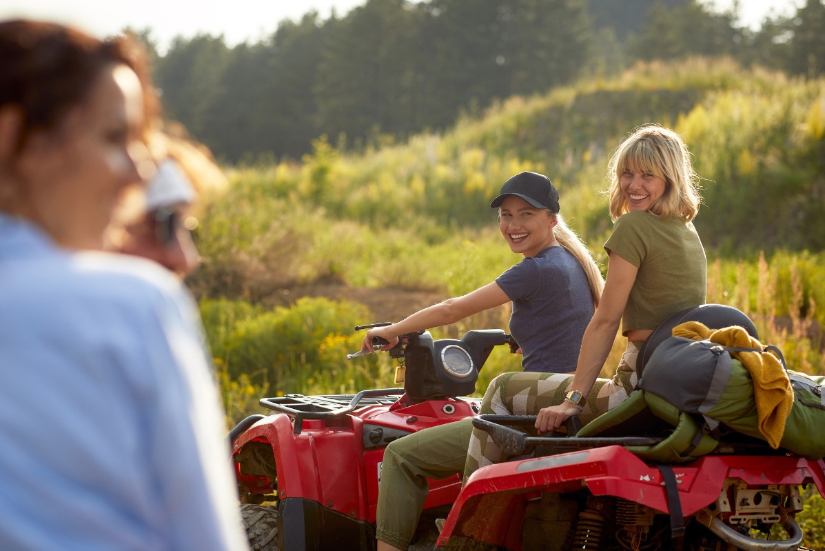 Dwie uśmiechnięte kobiety siedzą na czerwonym quadzie z plecakiem na bagażniku, w tle łąka i las, lekki efekt bokeh na pierwszym planie.